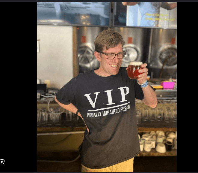 A Brewability team member smiling and holding a glass of amber beer in front of the color-coded tap wall.