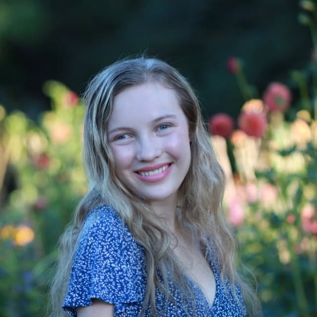 Alannah smiling outdoors in a blue and white patterned dress, with pink dahlias in bloom behind her.
