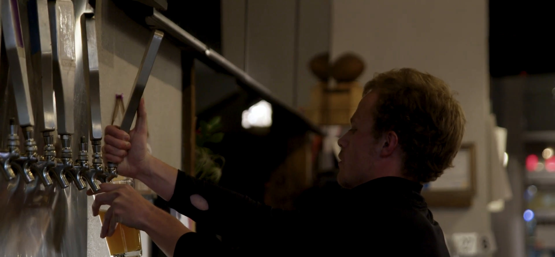 Avery reaching up to pull a tap handle, filling a glass of beer behind the Brewability bar.