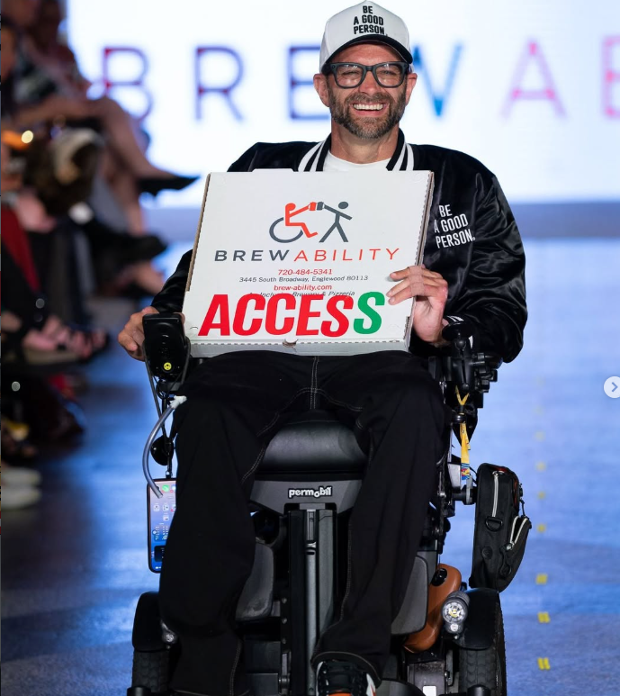 Jarod smiling as he holds a Brewability pizza box marked Access during a runway event.