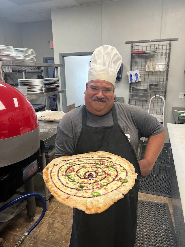 Jorge smiling in the Brewability kitchen while holding a freshly made pizza.