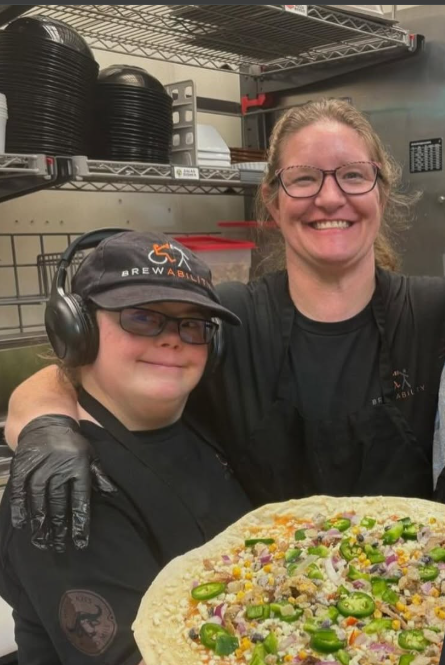 Mariah in a Brewability cap and headphones, standing with a coworker behind a large pizza in the kitchen.