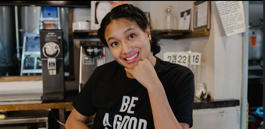 Miranda smiling beside the coffee equipment at Brewability in a black Be a Good Person shirt.