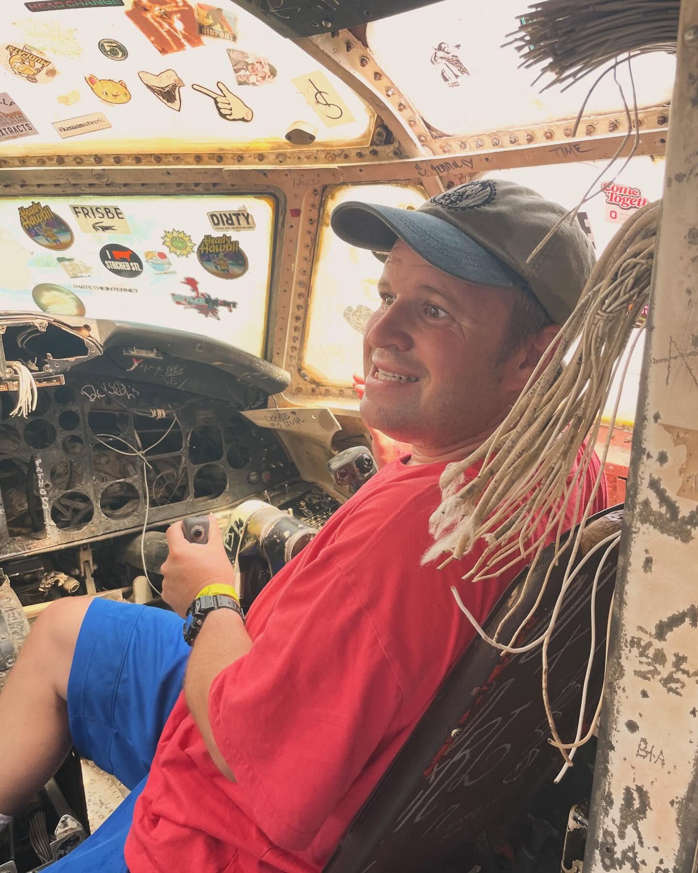 Nick smiling from the pilot's seat of an aircraft, wearing a red shirt and baseball cap.