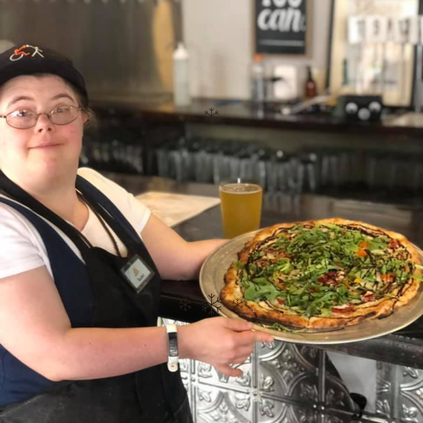 Rachel at the Brewability bar in her apron and name badge, holding out a large pizza.