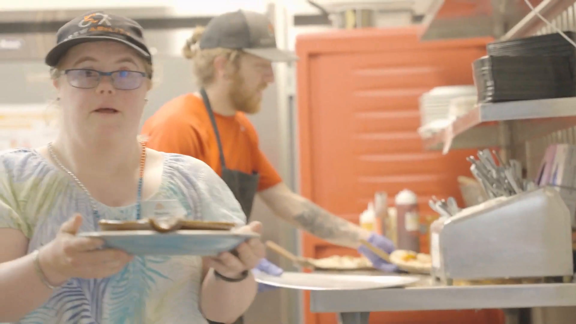 Rachel in her Brewability cap carrying a plate out from the pizza pass while a colleague works the oven behind her.